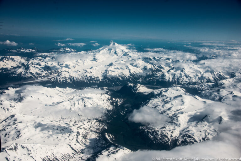 A distant pointed summit rises beyond layered snowy ridges and cloud-shadowed valleys.