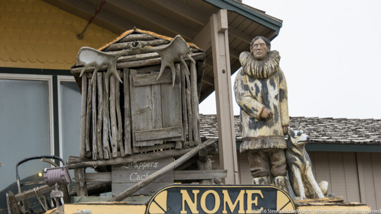 A Nome sign sits before a rustic wooden structure flanked by carved figures and hanging objects.