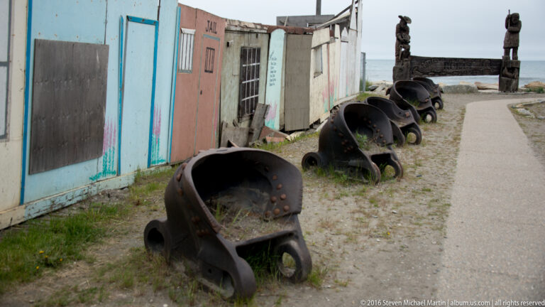 Rusty scoop-shaped metal containers line a gravel walkway beside weathered town buildings.