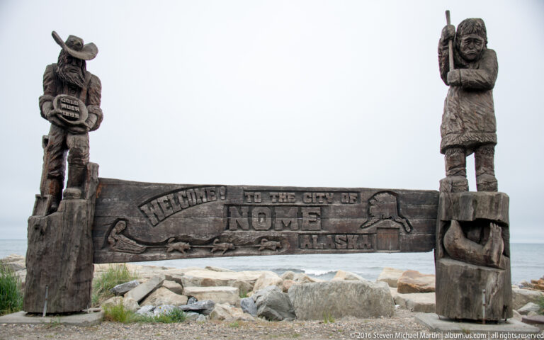 A carved wooden gateway welcomes visitors to Nome, Alaska, above rocks beside the sea.