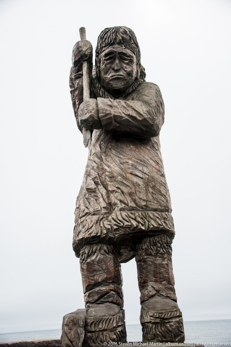 A carved figure in a parka and boots holds a long narrow object upright against the sky.