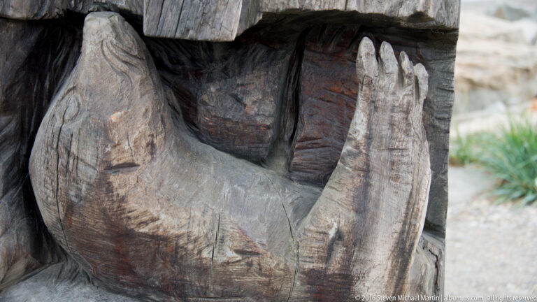 Close view of carved hands resting over a rounded wooden form, with a narrow strip of grass at right.