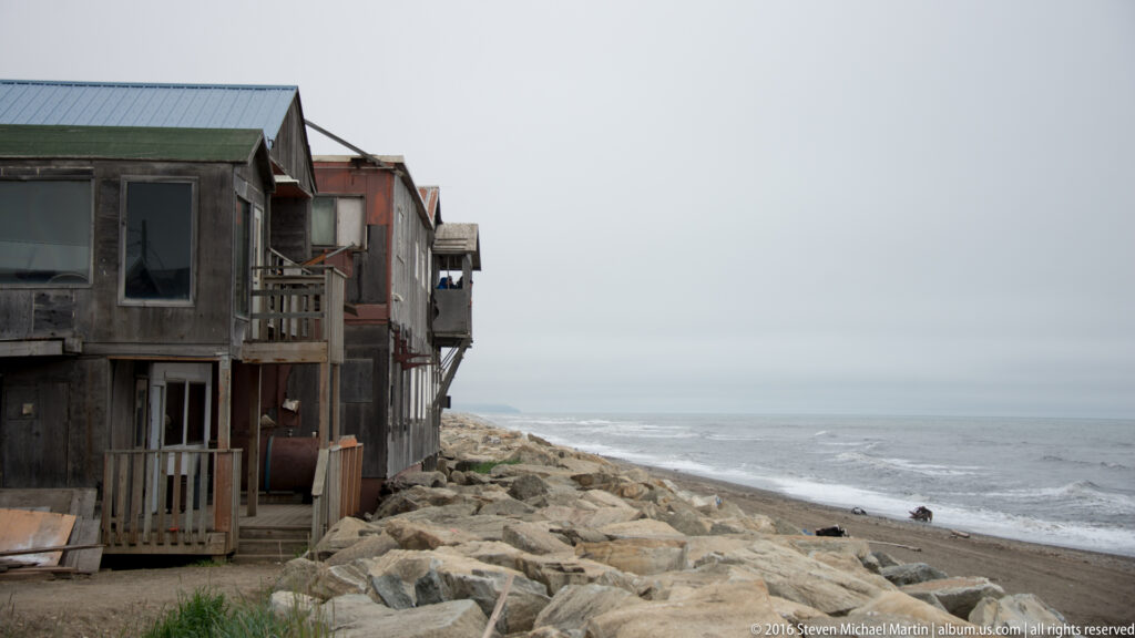 Weathered houses with porches stand beside a rocky beach and gray sea.