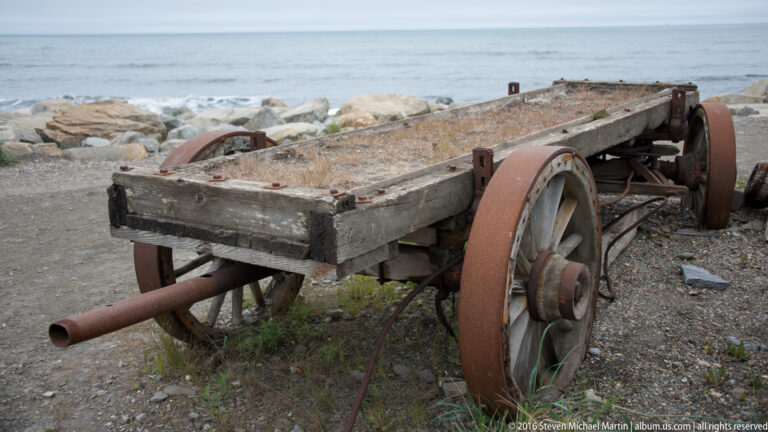 A weathered wooden cart with large rusted wheels and a projecting metal shaft rests beside the water.