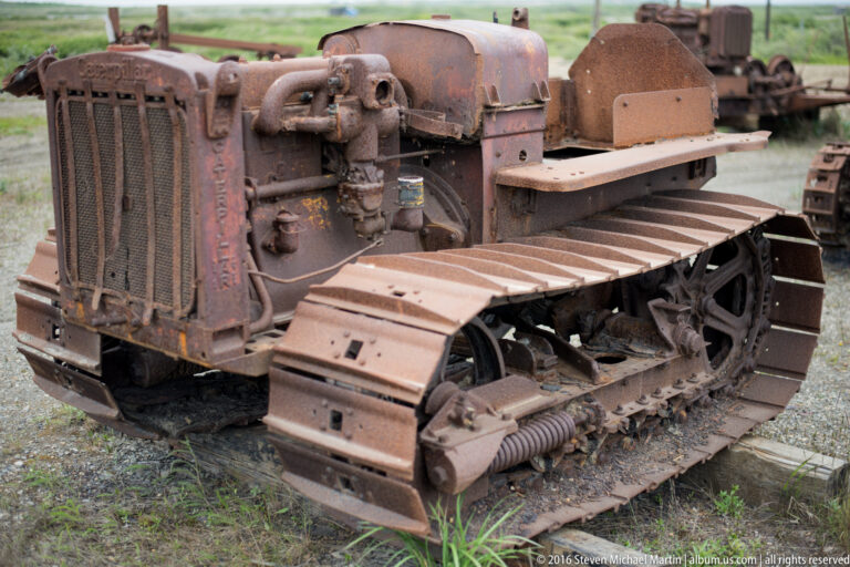 A rusted crawler tractor with an exposed engine, open seat, and broad metal tracks sits outdoors.