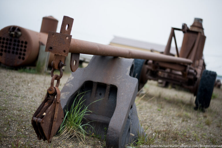 A heavy chain hangs from a rusted bracket beside a curved housing and large pipe.