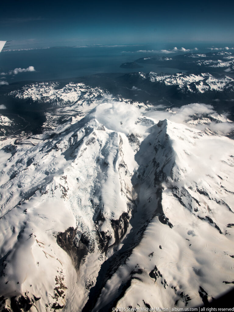 Cloud shadow darkens a foreground ridge while sunlit snowfields and peaks extend toward the horizon.