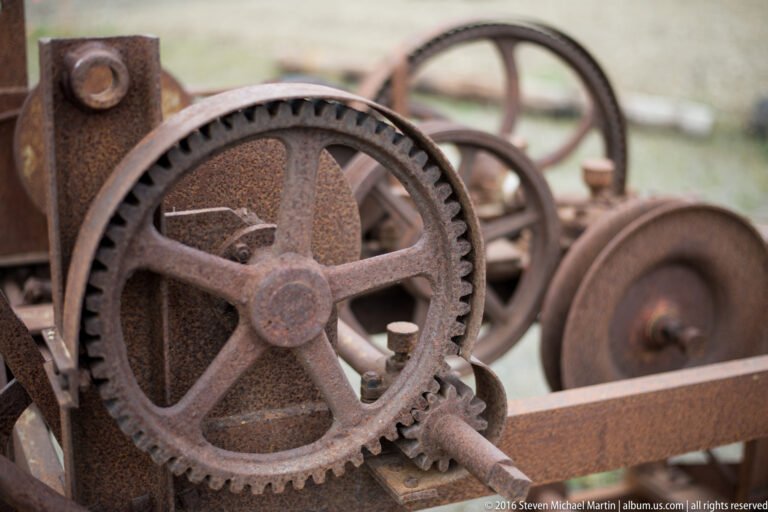 A large rusted wheel with broad spokes and toothed rim overlaps smaller gears and shafts.