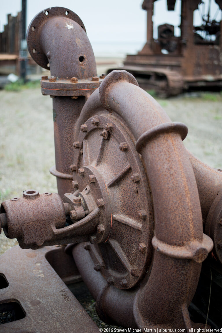 A rusted rounded housing with a bolted circular plate sits beside a curved pipe.
