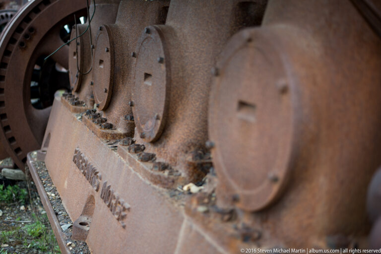 Round covers and heavy bolts line a rusted beam beneath gears and brackets.