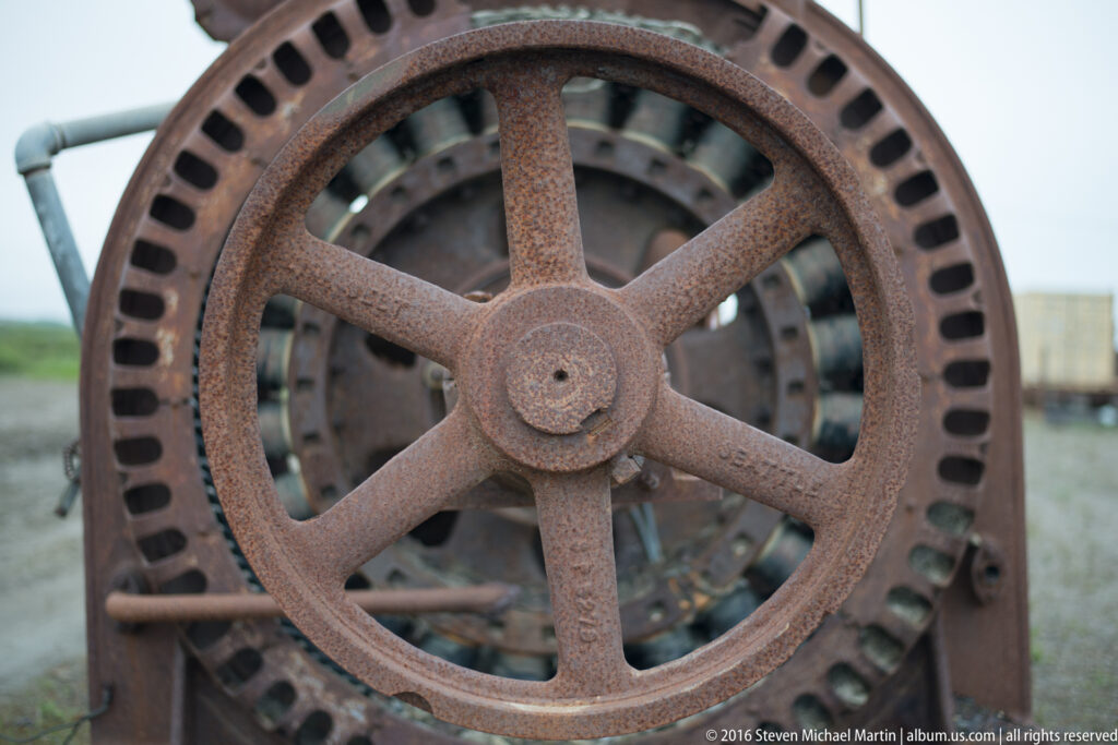 A large six-spoke rusted wheel sits before a perforated circular machine housing.