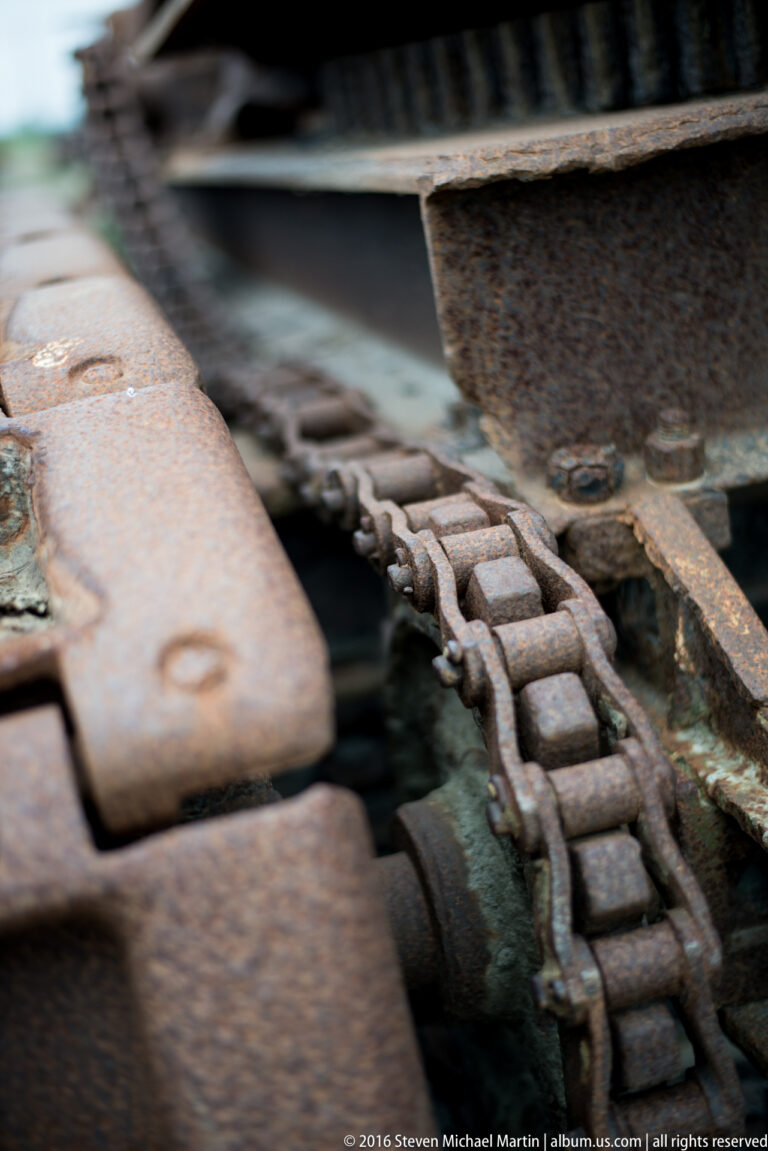 A heavy rusted metal track with linked plates and pins runs diagonally between steel components.