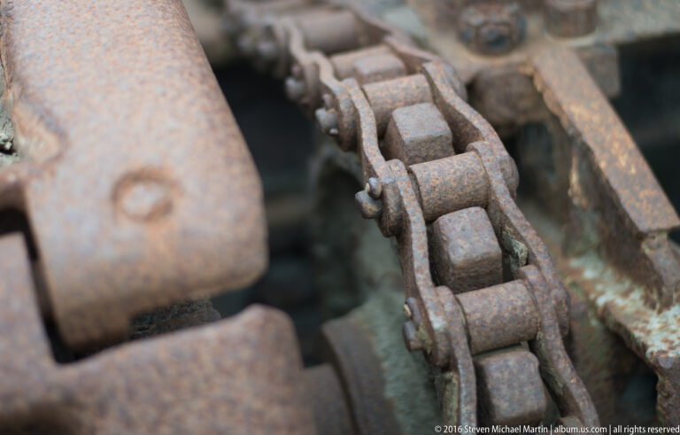 Corroded track links and pins recede between a flat metal plate and a curved steel rail.