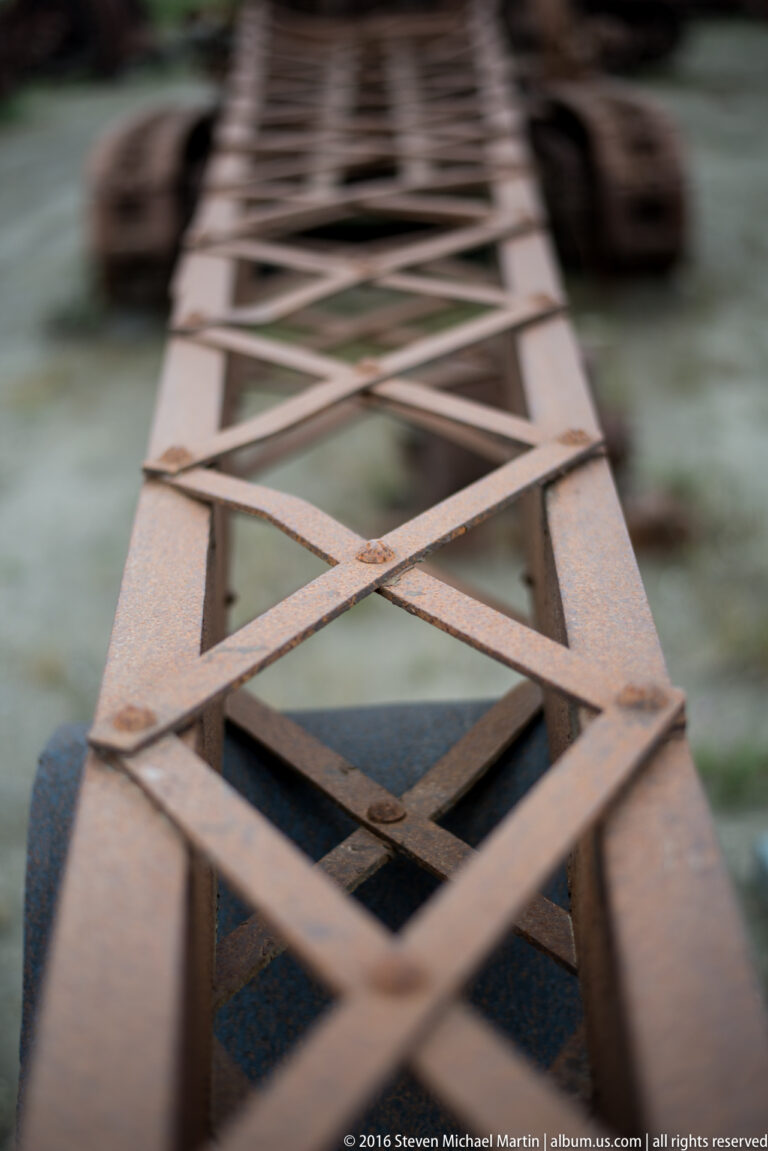 Diagonal metal braces form a narrowing lattice above a rusted structure, with the far end out of focus.