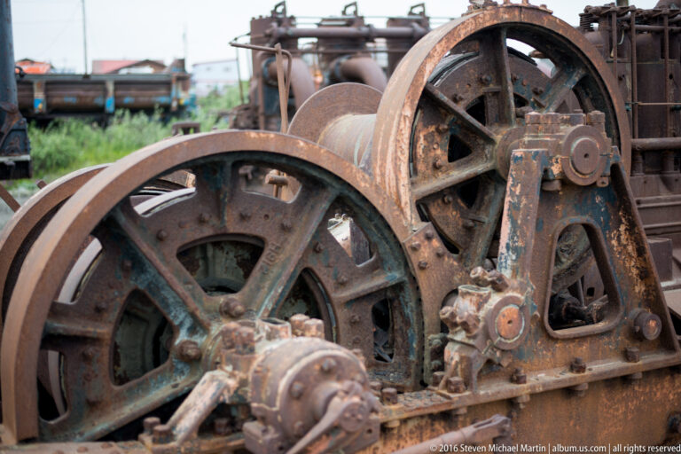 Two large rusted spoked wheels sit among axles, gears, and a cylindrical drum in an old machine.