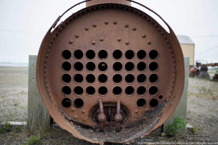 A rusted circular plate with rows of round openings and two fittings faces the camera.
