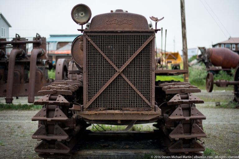 A rusted tracked machine with a square grille and cross braces stands among other equipment.