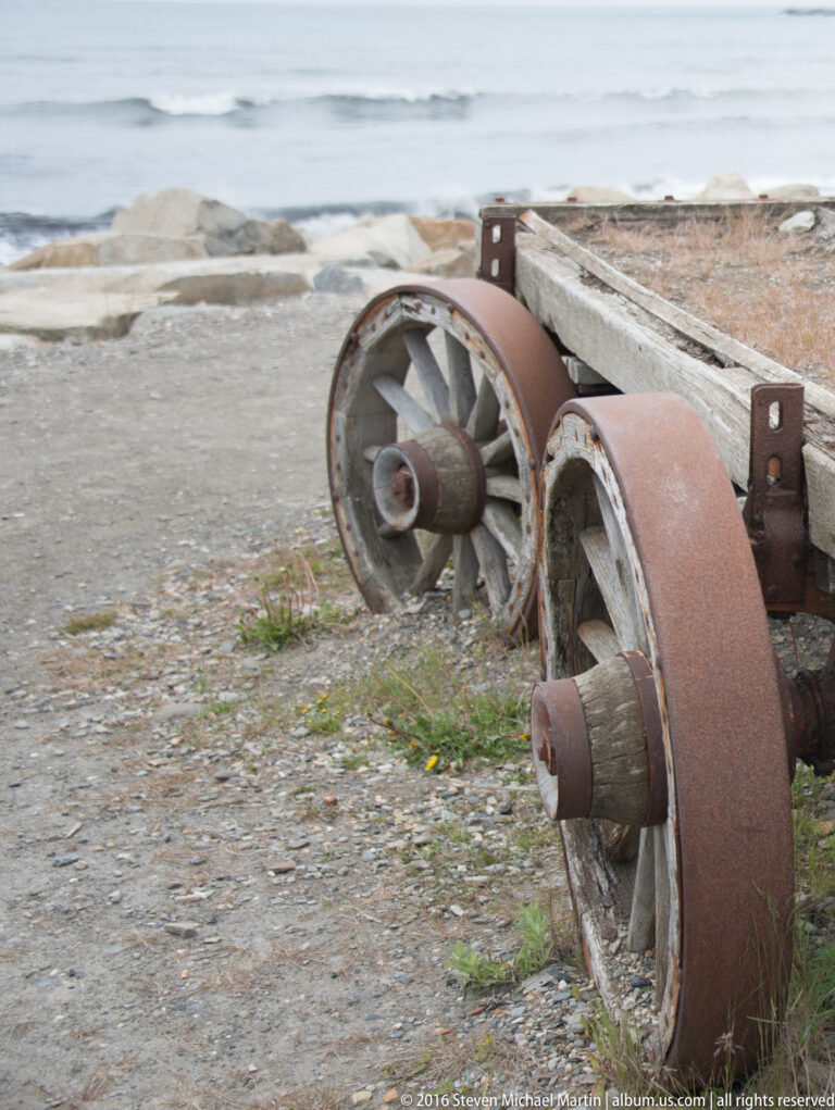 Two rusted iron wheels and an axle rest on a pebbled beach beside the water.