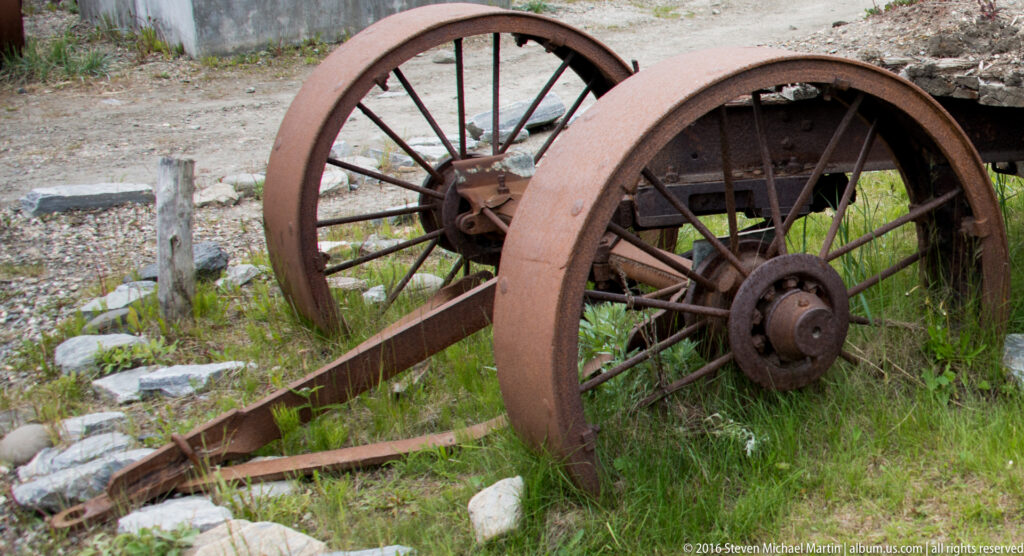 Two rusted spoked wheels sit in a low frame among grass and stones.