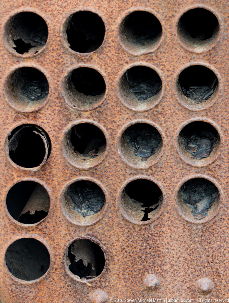 Staggered rows of large round holes puncture a rusted metal plate, with several openings damaged.