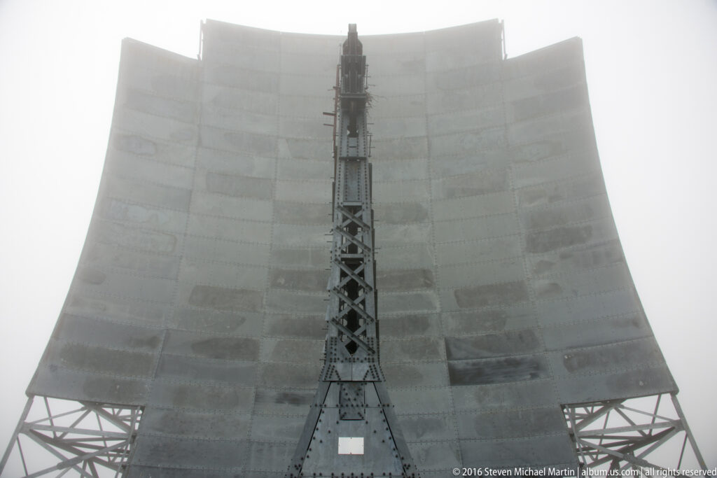 A tall lattice tower divides a broad segmented reflector antenna in fog.