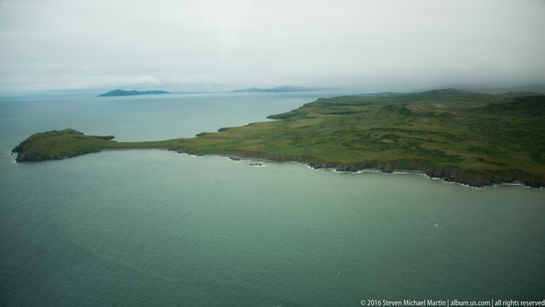 An aerial view shows green headlands, a sheltered inlet, pale water, and a distant island beneath low clouds.