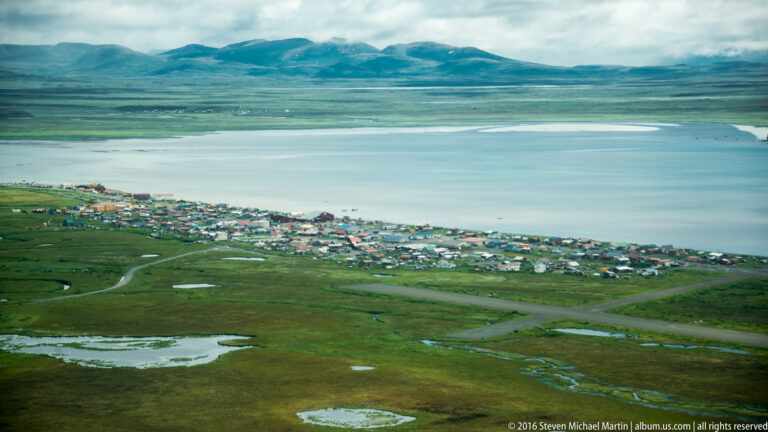 An aerial view shows a coastal village between a broad bay, green wetlands, and distant mountains.