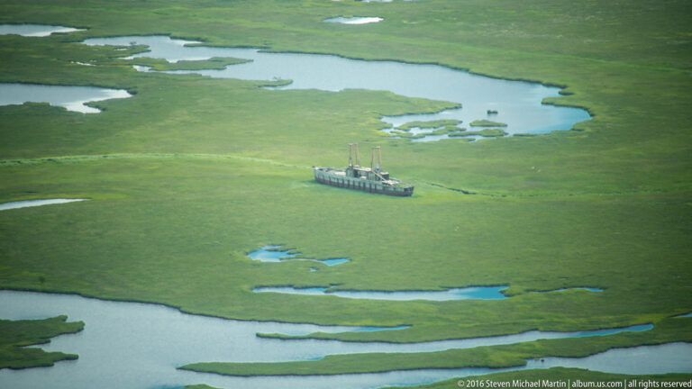 A small vessel with upright deck equipment sits amid green marshland and winding blue channels.