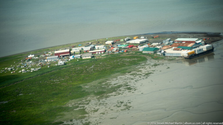 An aerial view shows clustered buildings and vehicles along an exposed muddy shoreline at low tide.