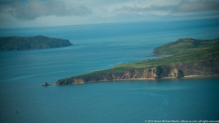 An aerial view looks across blue water toward a steep, layered headland and a green island beyond.