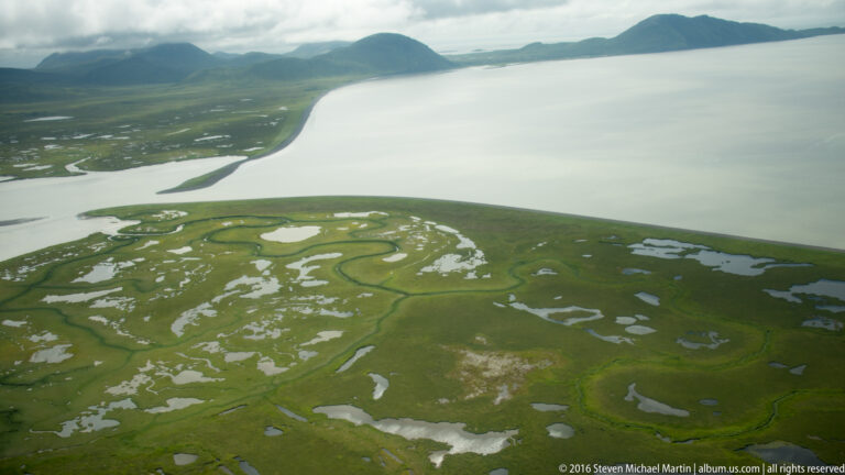 A broad bay curves past green marshland patterned with winding channels and shallow pools.
