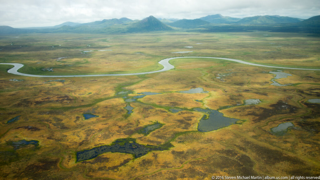 An expansive green and ochre wetland with scattered pools and a winding river stretches toward distant mountains.