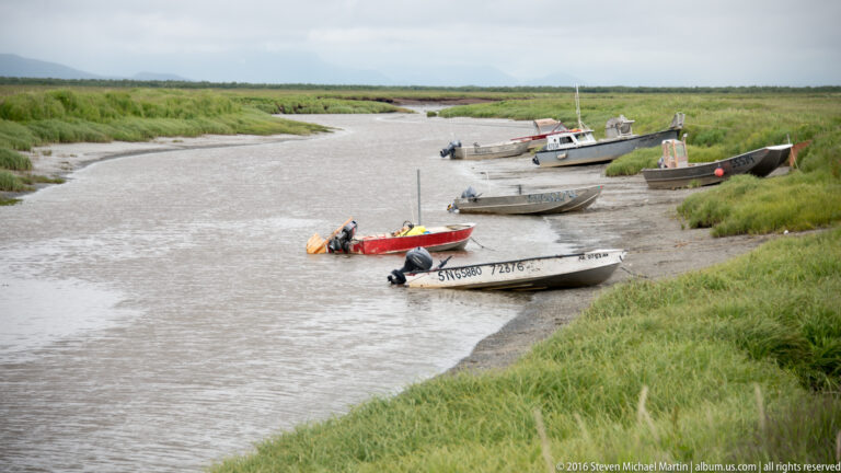 Small boats, including a red one in front, rest in a shallow channel bordered by green marsh banks.