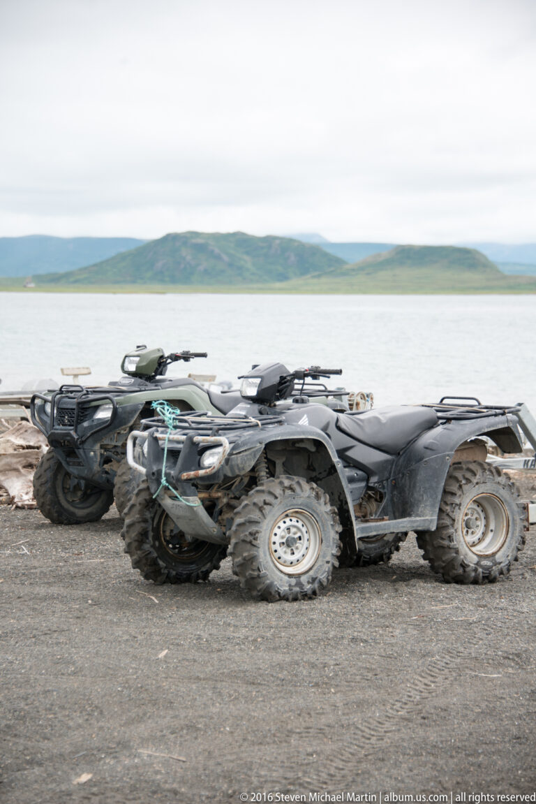 Several all-terrain vehicles stand on a gravel shore above calm water and low green hills.
