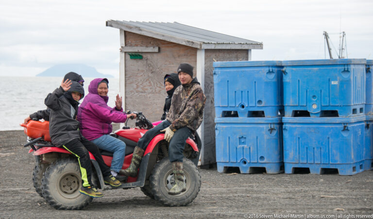 Three people sit on a red ATV beside a shed and stacked blue containers, one raising a hand toward the camera.