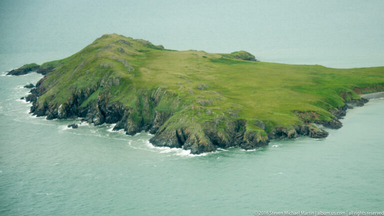 A grassy peninsula with steep rocky sides projects into blue-green water edged by white surf.