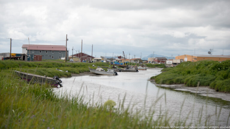 Several boats line a narrow channel through a village, bordered by tall grass and low buildings.