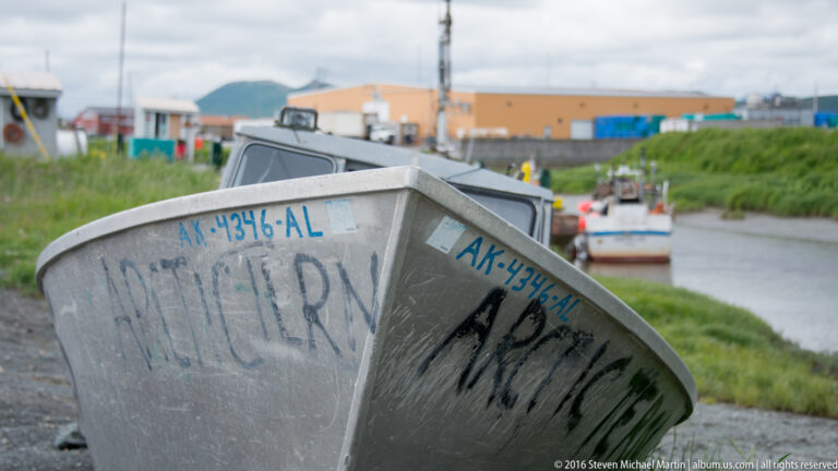 A small aluminum skiff with blue registration and dark handwritten markings sits beside a village channel.