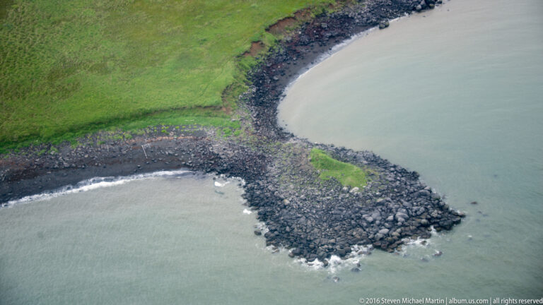 A green bank curves between two inlets and ends in a dark rocky spit beside pale shallow water.