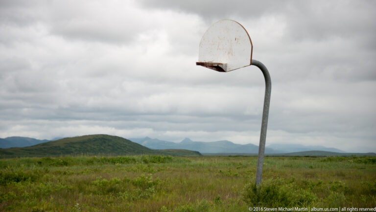 A weathered basketball hoop stands in a grassy wetland, with low hills and distant mountains beneath cloudy skies.
