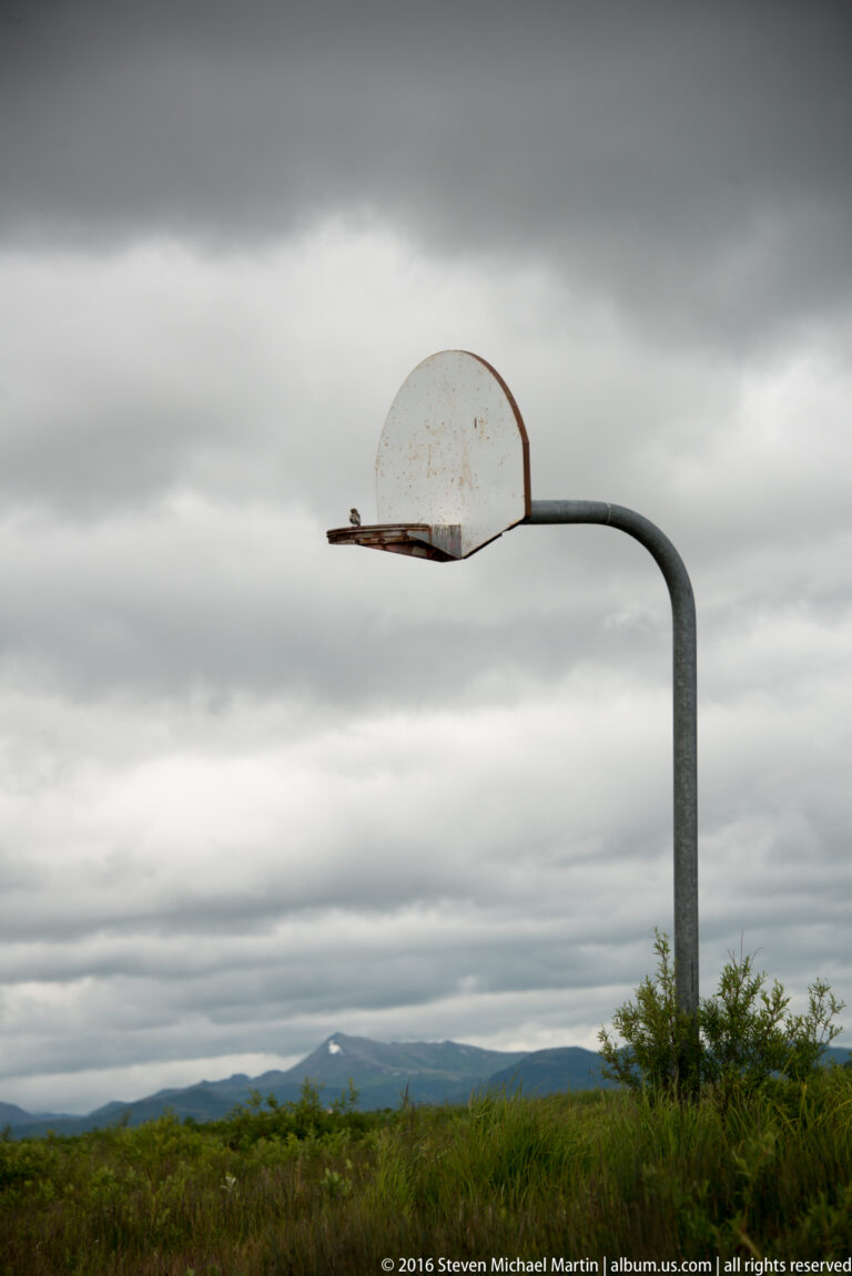 A curved metal pole supports a weathered basketball backboard above green vegetation, with a mountain ridge in the distance.