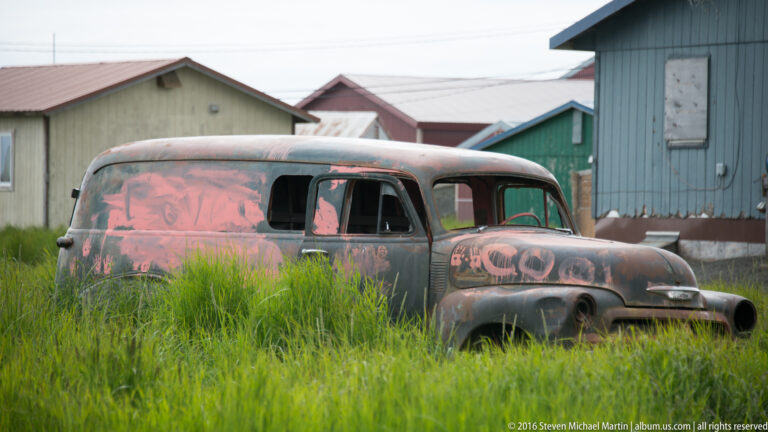 A rusted old pickup truck rests in tall grass between simple buildings.