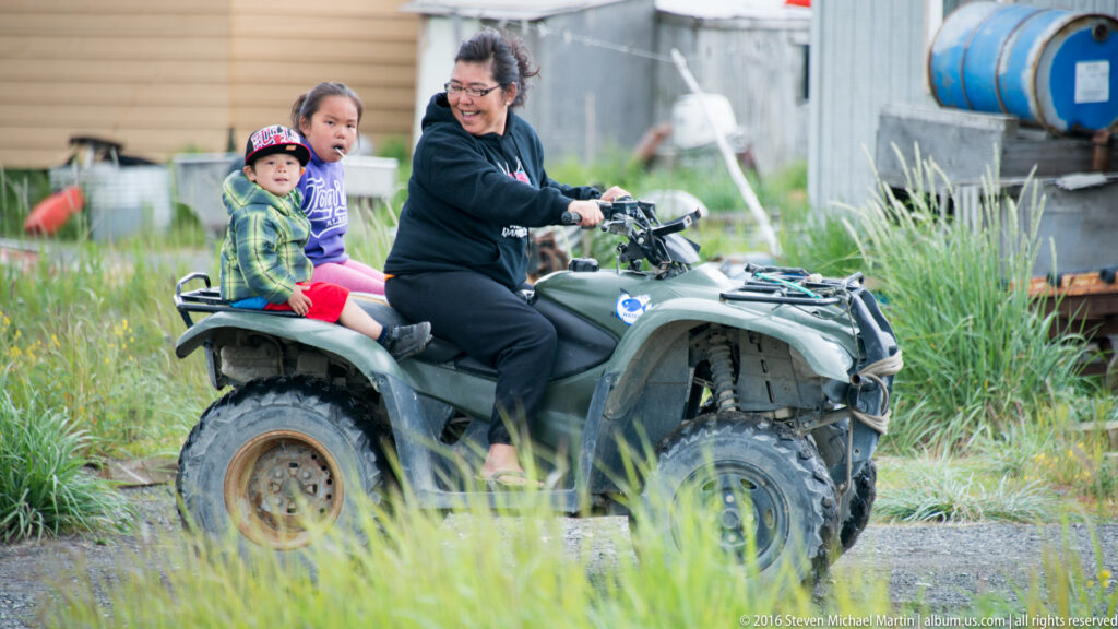 A woman drives an all-terrain vehicle with two children seated behind and beside her on a grassy village path.