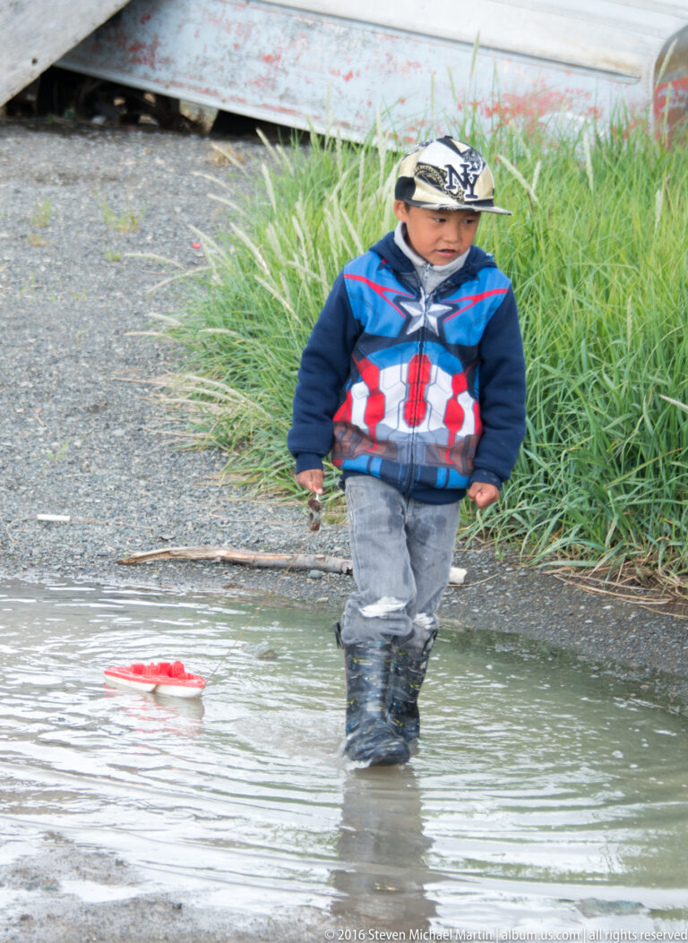 A child in a patterned cap wades through shallow water beside grass, with a small red object floating nearby.