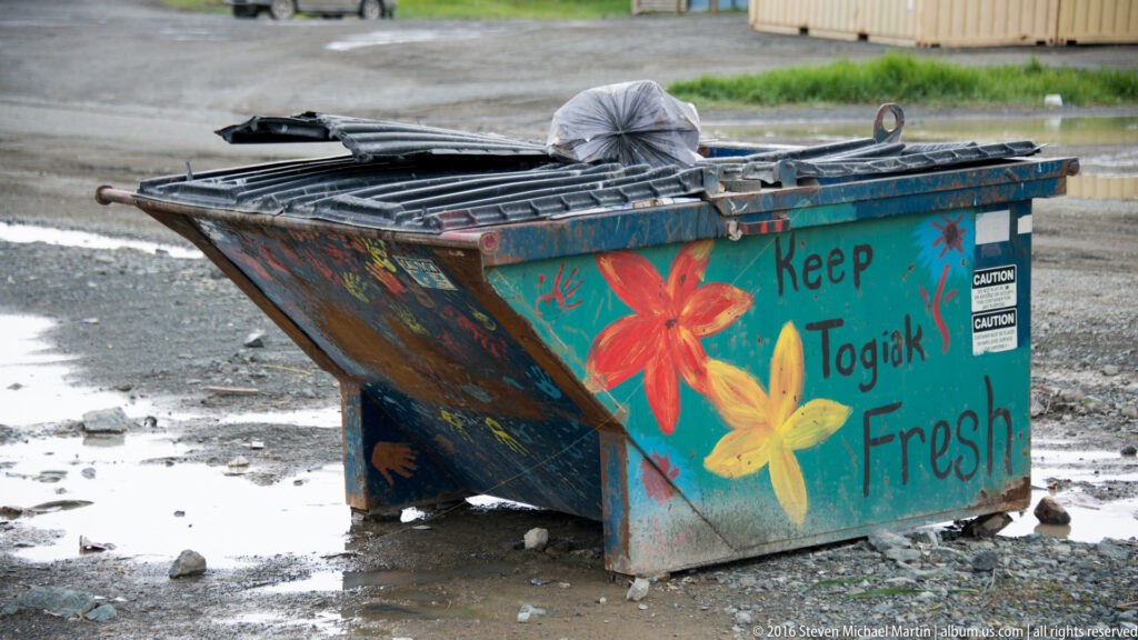 A blue dumpster painted with flowers and the words “Keep Togiak Fresh” stands on wet gravel.