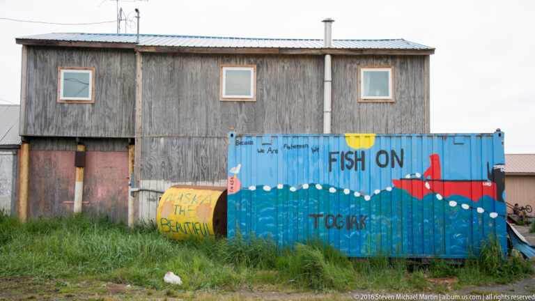 A blue container painted with a red fish and the words “FISH ON” and “TOGIAK” stands before a wooden building.