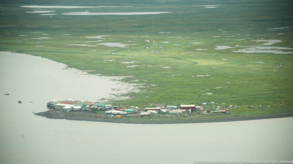 A small settlement lines the shore beside a broad waterway and expansive green wetland.