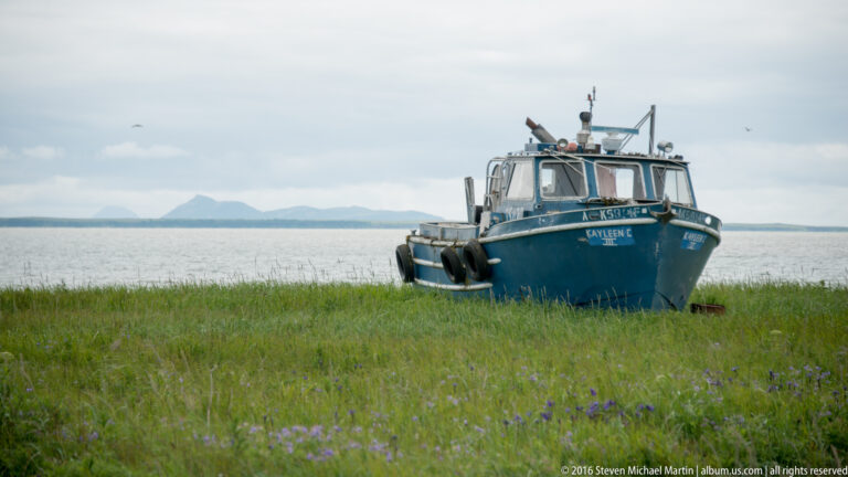 A blue fishing boat named Kayleen C sits at a grassy shoreline, with water and distant mountains behind it.