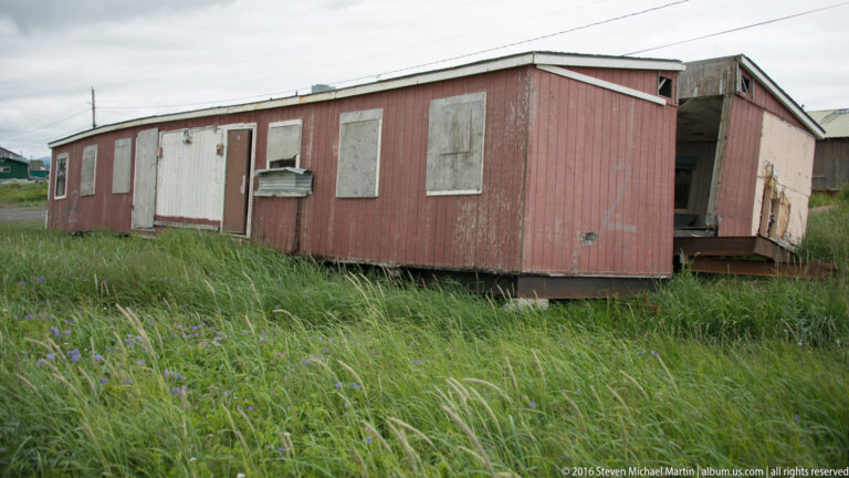 A weathered red outbuilding with boarded windows and an open end stands in tall grass.