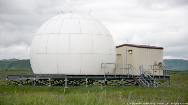 A large white spherical dome stands on a raised metal platform beside a small building in open tundra.